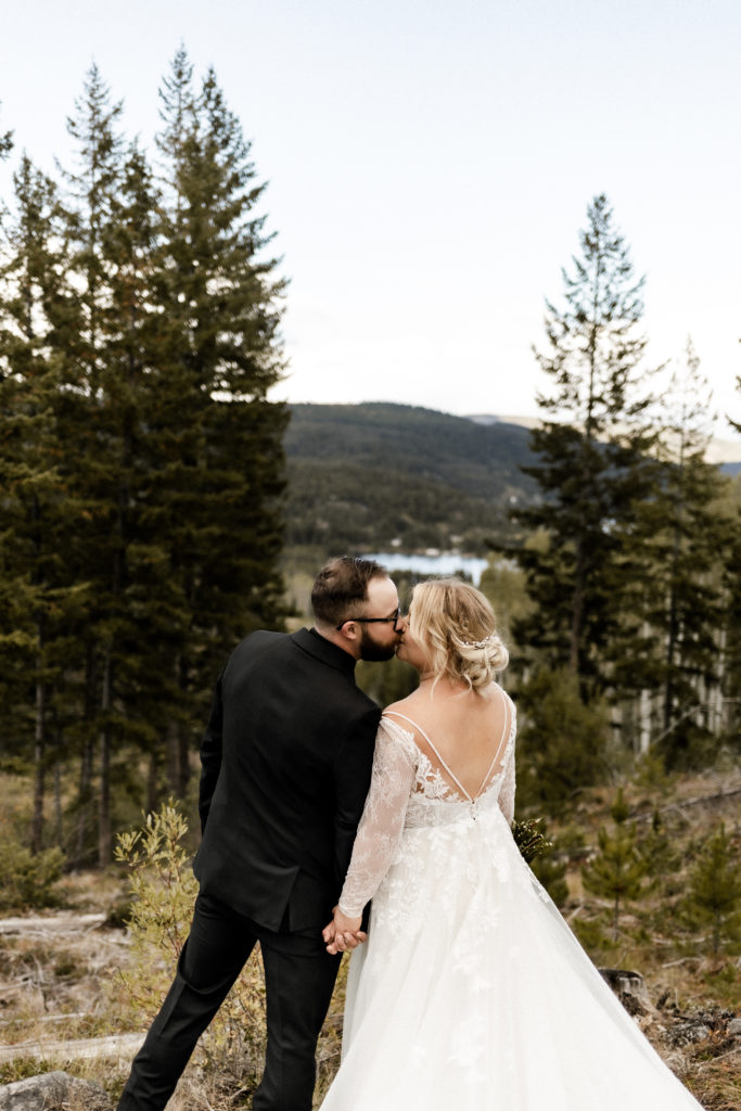 A Caucasian couple stands atop a hill, overlooking a tranquil blue lake, embraced in a kiss. Surrounding them are lush green trees, and their backs are turned toward the camera, symbolizing intimacy and connection amidst nature's beauty.