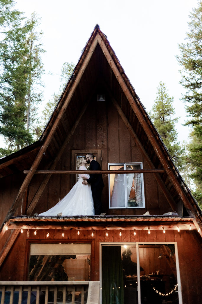 A Caucasian couple embraces on the deck of their A-frame cabin, exchanging a kiss. The bride, in a white lace dress, leans into the groom, dressed in black. Fairy lights adorn the cabin, casting a warm glow.