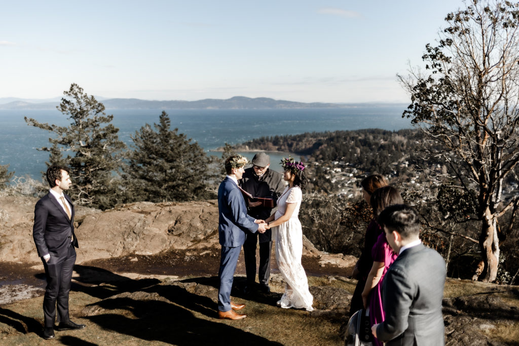An intimate elopement captured on top of a mountain with a breathtaking ocean view. The couple stands hand in hand during their ceremony, radiating love and joy. The partner on the left wears a simple white dress, while the one on the right is adorned in a navy suit. Both wear delicate flower crowns, adding a touch of natural beauty to their special moment.
