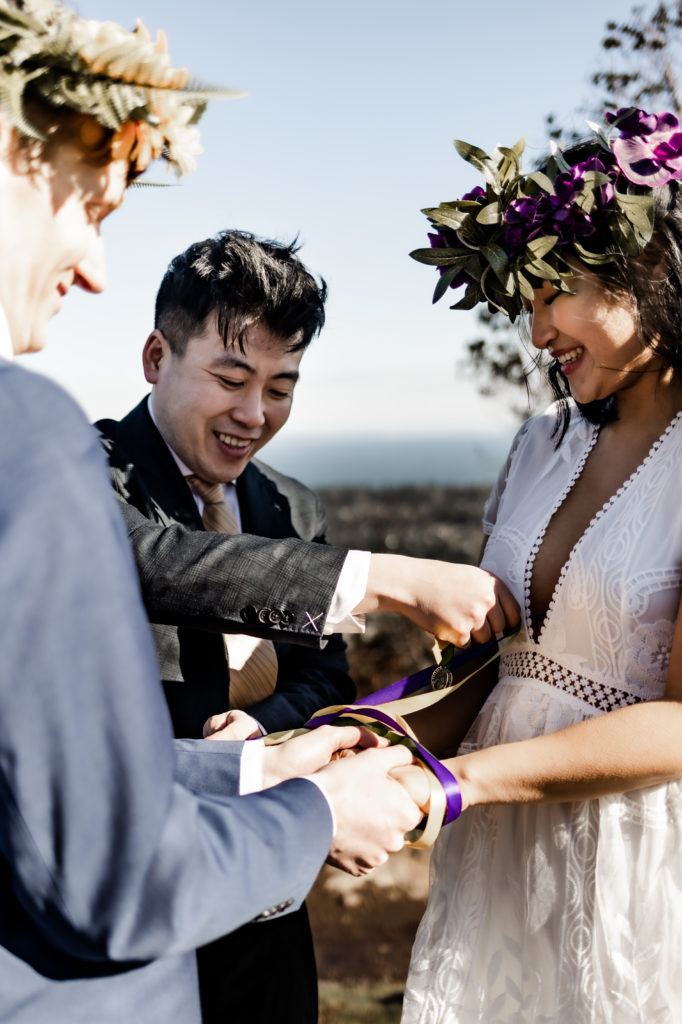 In an intimate ceremony atop the mountain, the couple, adorned in a navy suit and a white dress, participate in a ribbon ritual. The close-up shot captures the intricate details as they delicately weave colorful ribbons, symbolizing the intertwining of their lives. Against the scenic backdrop of the ocean, the couple's focused expressions reflect the depth and significance of this heartfelt ceremony, creating a moment of unity and connection on their special day.