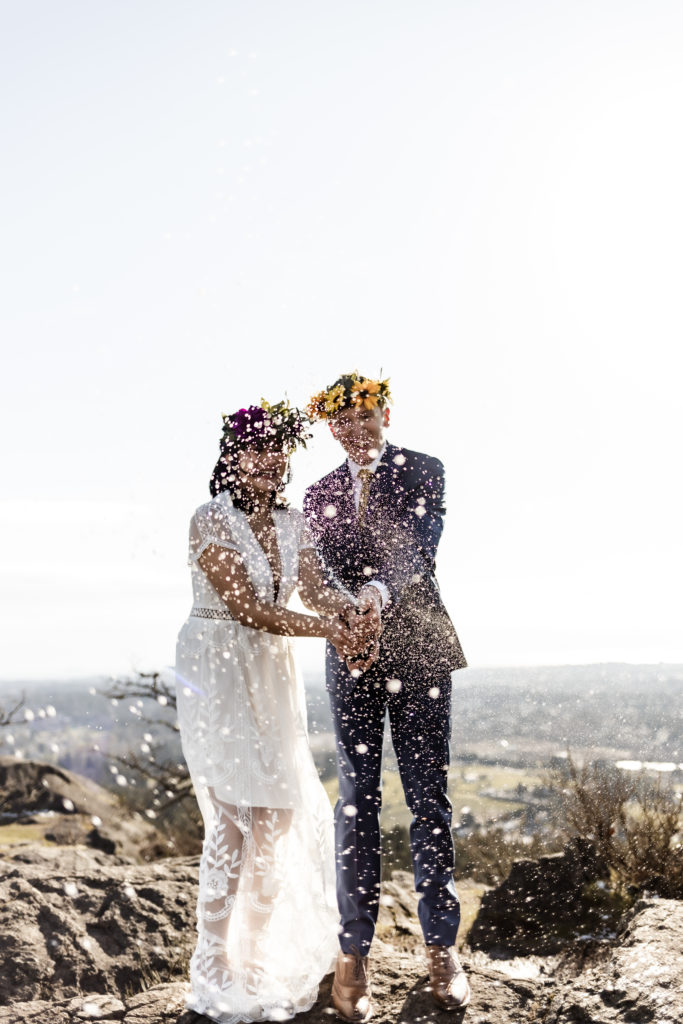 Amidst the breathtaking mountain and ocean vista, the couple, donned in a navy suit and a white dress, jointly wields a champagne bottle. The camera focuses on the effervescent spray of champagne, capturing a whimsical moment of shared celebration. Laughter fills the air as the couple revels in this playful act, creating a dynamic and joyous scene against the backdrop of nature's grandeur.