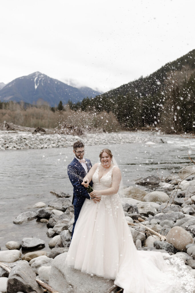 At a beautiful elopement in front of Chilliwack River. A couple stands before the gorgeous mountain view, spraying a bottle of champagne at the camera. One partner wears a large, poofy white wedding dress, while the other partner wears a navy suit with a pink bowtie.