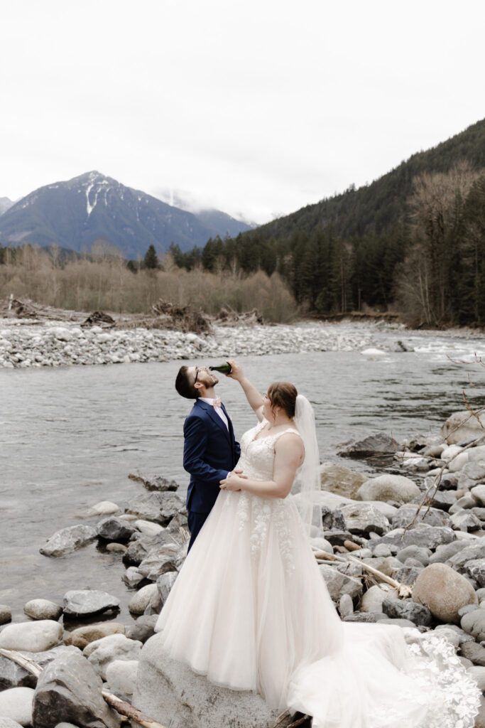 At a beautiful elopement in front of Chilliwack River. A couple stands before the gorgeous mountain view. One partner wears a large, poofy white wedding dress, while the other partner wears a navy suit with a pink bowtie. The partner wearing the white dress pours the remaining champagne into the mouth of the partner wearing a suit after they complete their champagne pop.