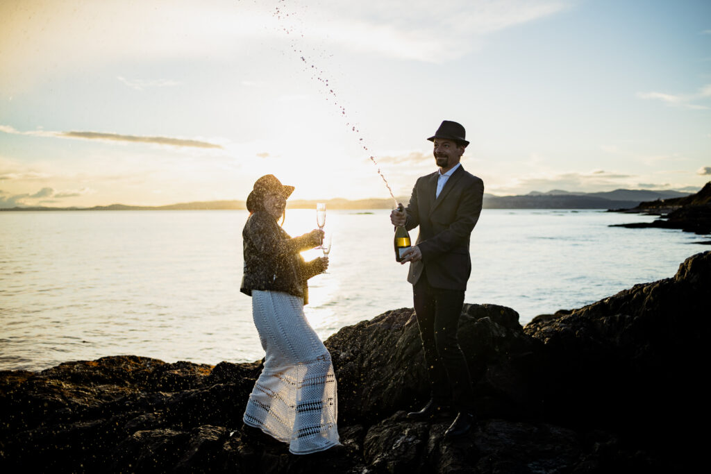Standing in front of a beautiful sunset is a couple doing a champagne pop. One partner is wearing a top hat and suit, and the other is wearing a leather jacket and white boho dress. The partner wearing the suit sprays the champagne into the air, creating a shower of liquid. The partner in the white dress leans away from the spray with a laugh, holding a champagne flute.