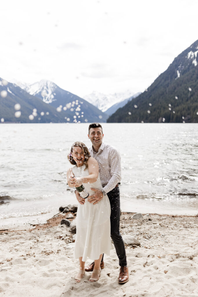 A couple stand together in front of the Chilliwack Lake mountains, spraying a bottle of champagne at the camera. One partner stands behind the other with his hands on her waist as the other partner sprays the bottle. The partner in the back wears a classy white patterned shirt with dress pants and shoes. The partner spraying the bottle wears a calf length white dress.