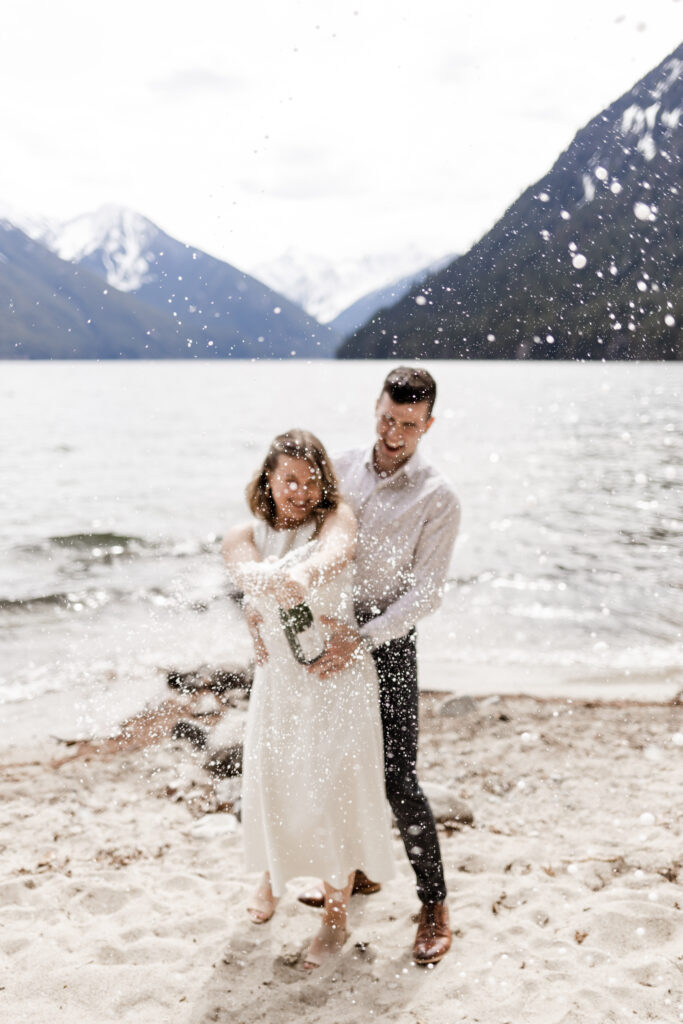 A couple stand together in front of the Chilliwack Lake mountains, spraying a bottle of champagne at the camera. One partner stands behind the other with his hands on her waist as the other partner sprays the bottle. The partner in the back wears a classy white patterned shirt with dress pants and shoes. The partner spraying the bottle wears a calf length white dress.
