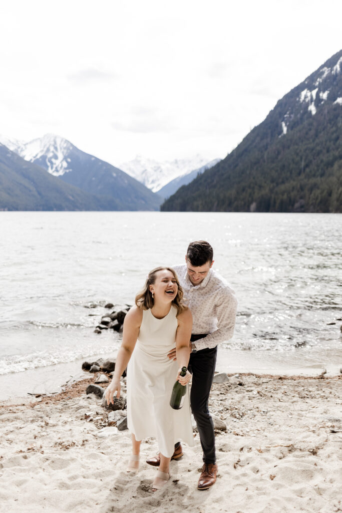 A couple shares a laugh immediately after their champagne pop in front of Chilliwack Lake. Both partners lean forward in laughter.