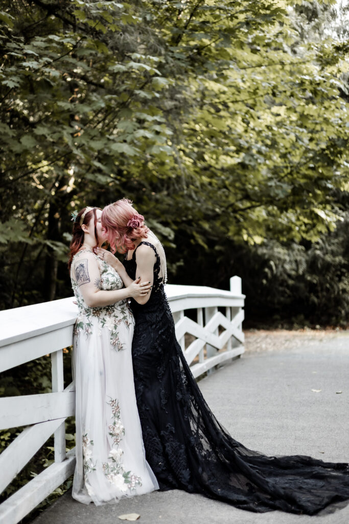 An LGBTQ+ caucasian couple share a kiss while leaning against a white railing on a bridge. One partner wears a black lace dress, and the other wears a white floral dress.