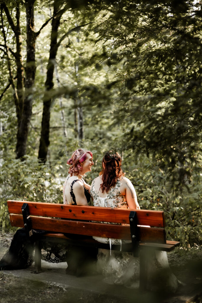 An LGBTQ+ caucasian couple share a laugh while sitting on a bench in the forest. One partner wears a black lace dress, and the other wears a white floral dress.