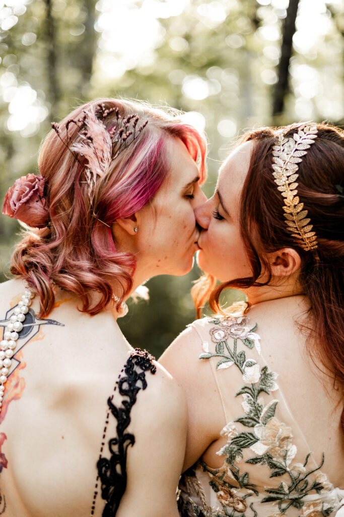 An up-close portrait of an LGBTQ+ caucasian couple sharing a kiss. One partner wears a black lace dress, and the other wears a white floral dress.