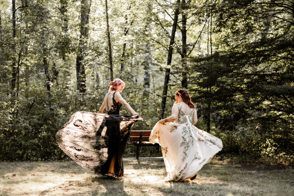 An LGBTQ+ caucasian couple twirl in their wedding dresses in the forest. One partner wears a black lace dress, and the other wears a white floral dress.