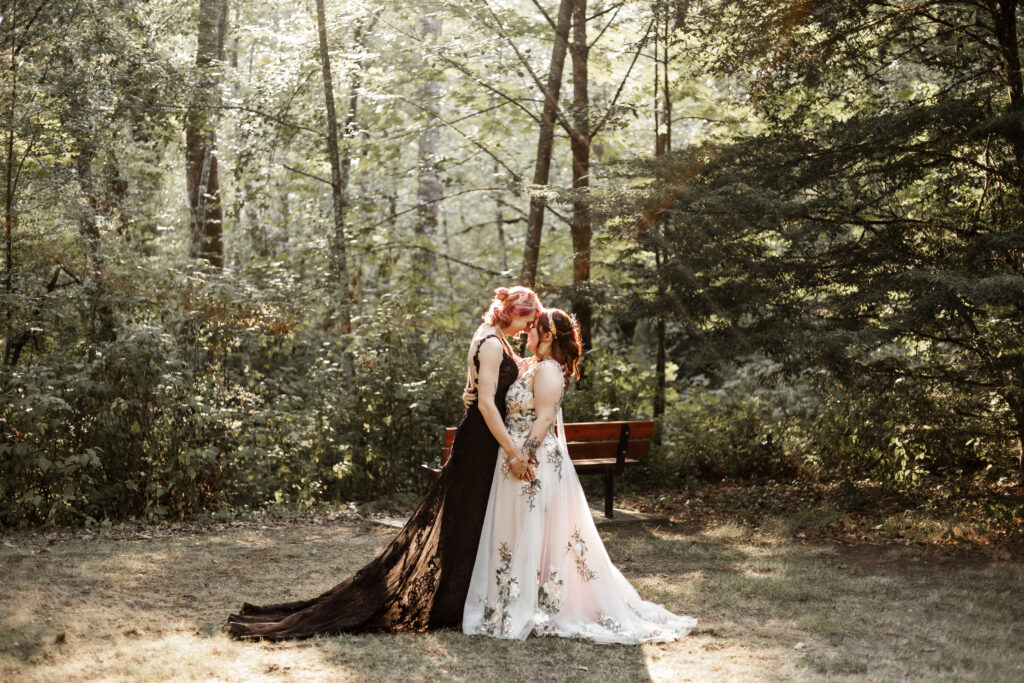 An intimate moment captured in a forest setting during an LGBTQ+ elopement. The couple, both Caucasian, touch noses in the forest as the sun shines through the trees. One partner wears a white flowery dress, while the other is dressed in a black lace gown.