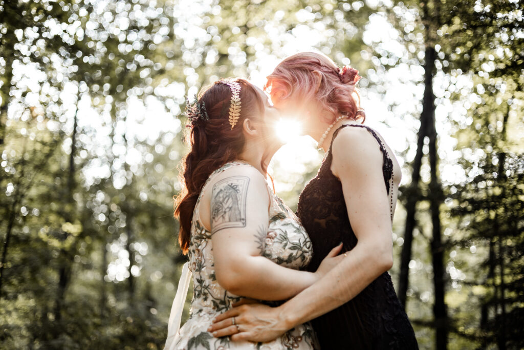 An intimate moment captured in a forest setting during an LGBTQ+ elopement. The couple, both Caucasian, share a kiss as the sun shines behind them. One partner wears a white flowery dress, while the other is dressed in a black lace gown.