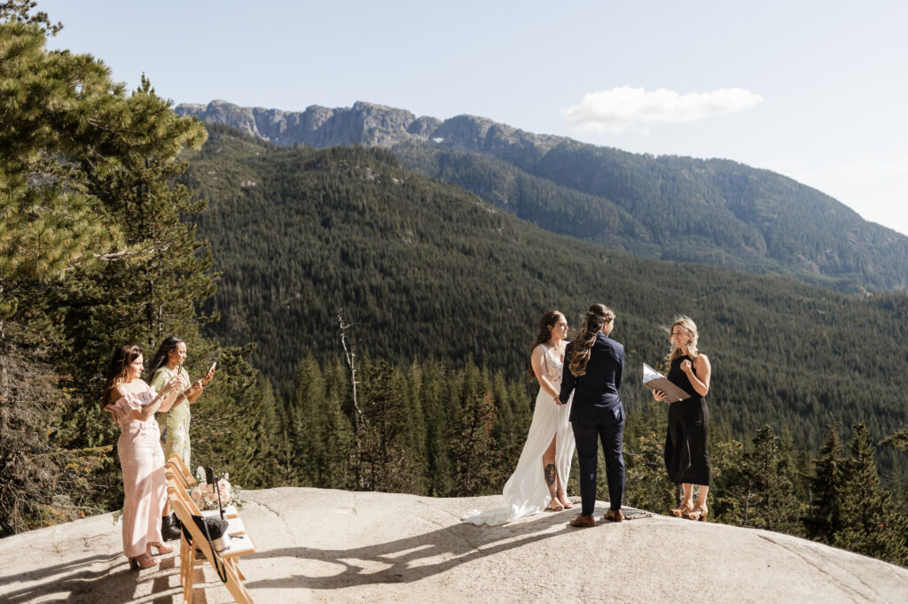 An LGBTQ+ elopement at the Sea to Sky Gondola in Vancouver. In the image, one partner wears a beautiful white dress, while the other dons a navy suit. They stand against a stunning backdrop of green mountains, while their family and friends watch from the side.