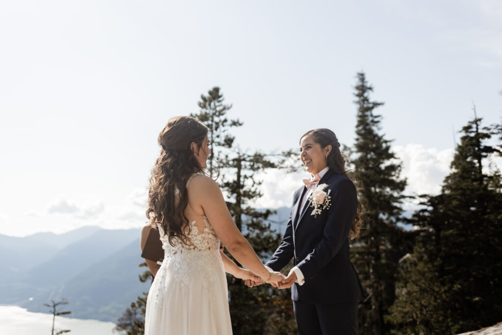 A breathtaking LGBTQ+ elopement at the Sea to Sky Gondola in Vancouver. In the image, one partner wears a beautiful white dress, while the other dons a navy suit. They stand against a stunning backdrop of majestic mountains and the vast ocean, exchanging heartfelt vows under the clear sky.
