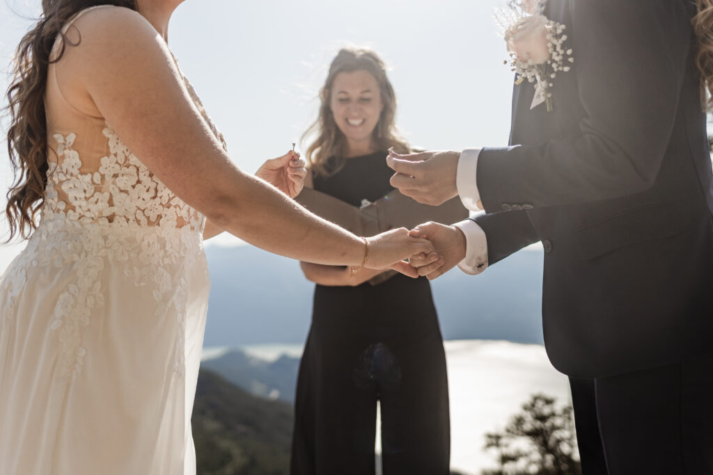 A breathtaking LGBTQ+ elopement at the Sea to Sky Gondola in Vancouver. In the image, one partner wears a beautiful white dress, while the other dons a navy suit. It's an up-close shot of the couple holding up their rings in preparation for the ring exchange.