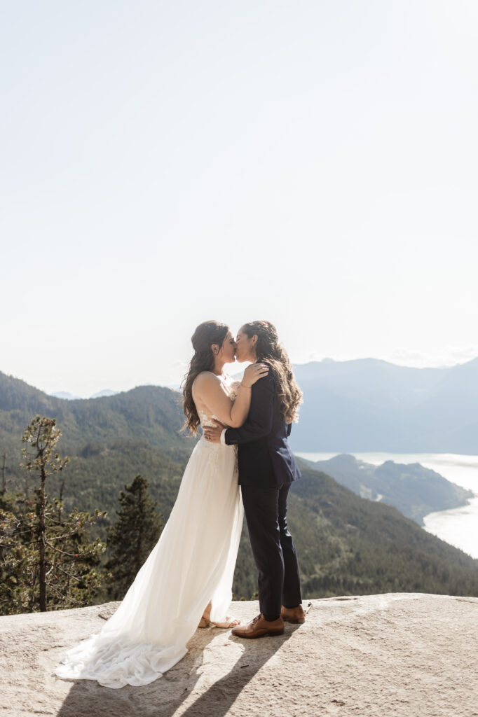 A breathtaking LGBTQ+ elopement at the Sea to Sky Gondola in Vancouver. In the image, one partner wears a beautiful white dress, while the other dons a navy suit. They stand against a stunning backdrop of majestic mountains and the vast ocean as they share their first kiss as a married couple.