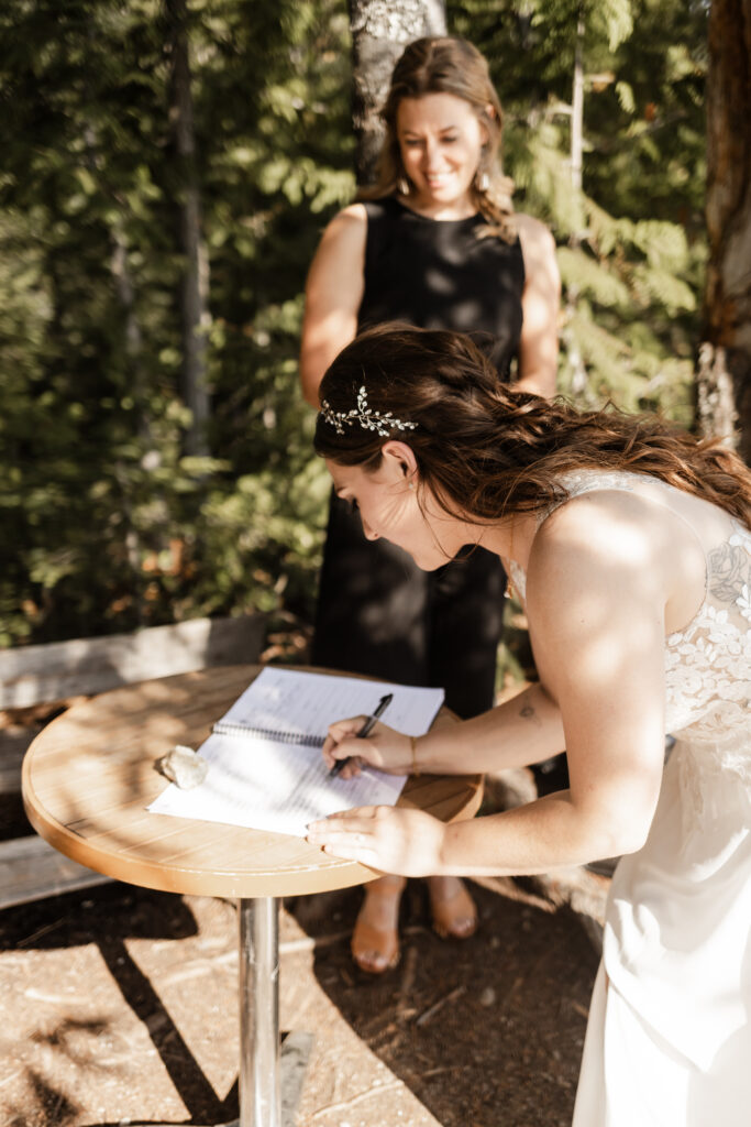 A breathtaking LGBTQ+ elopement at the Sea to Sky Gondola in Vancouver. In the image, one partner leans over a table to sign the marriage license as the officiant watches from behind.
