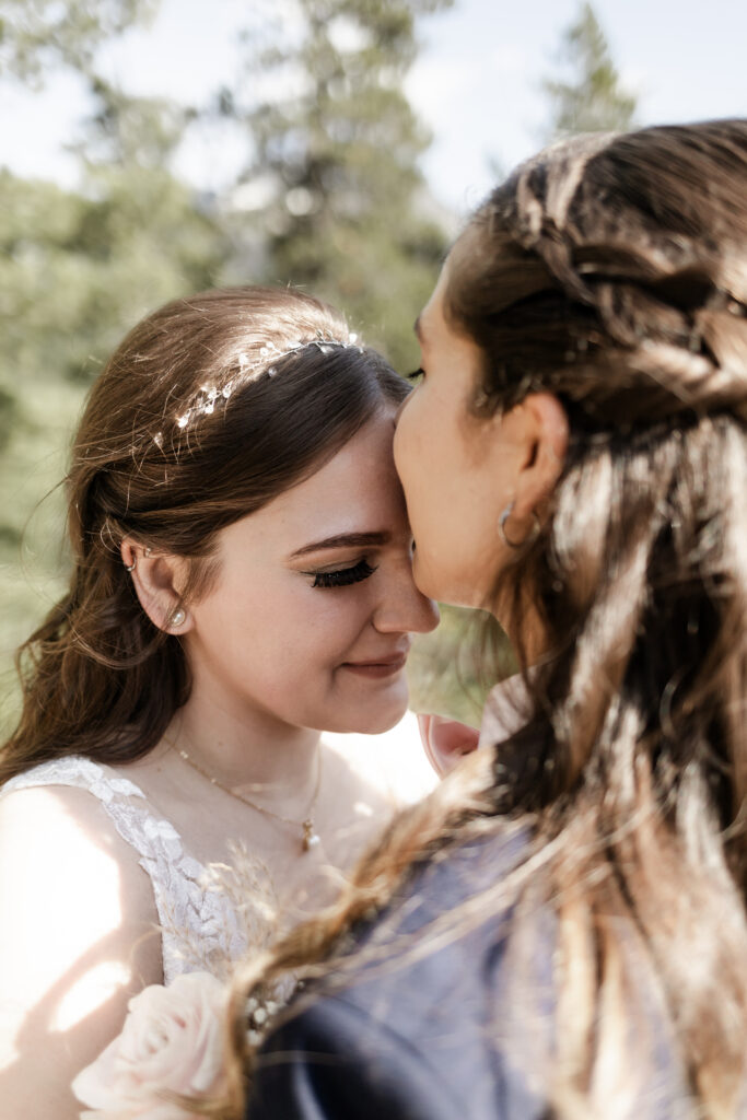 A breathtaking LGBTQ+ elopement at the Sea to Sky Gondola in Vancouver. In the image, one partner wears a beautiful white dress, while the other dons a navy suit. It's an up-close shot of the partner in the dress as the other partner kisses her forehead.