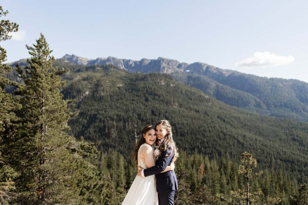 A breathtaking LGBTQ+ elopement at the Sea to Sky Gondola in Vancouver. In the image, one partner wears a beautiful white dress, while the other dons a navy suit. The couple shares an embrace in front of the mountain view.