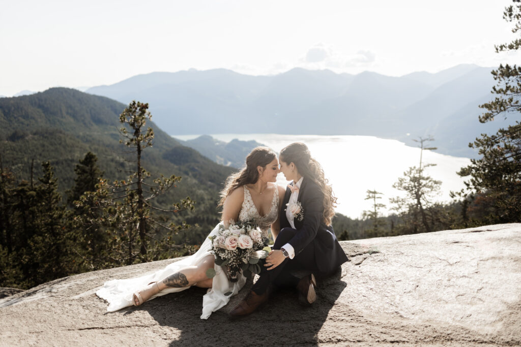 A breathtaking LGBTQ+ elopement at the Sea to Sky Gondola in Vancouver. In the image, one partner wears a beautiful white dress, while the other dons a navy suit. They sit against a stunning backdrop of majestic mountains and the vast ocean, touching their noses together.
