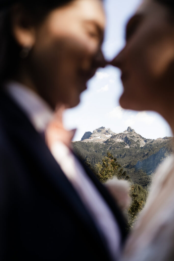 An intimate moment captured as the LGBTQ+ couple tenderly touches noses, their love taking center stage in this close-up shot. The camera skillfully shifts focus, highlighting the blurred outlines of the couple against a majestic backdrop of mountains, creating a beautifully framed image that symbolizes their connection against the breathtaking natural scenery.
