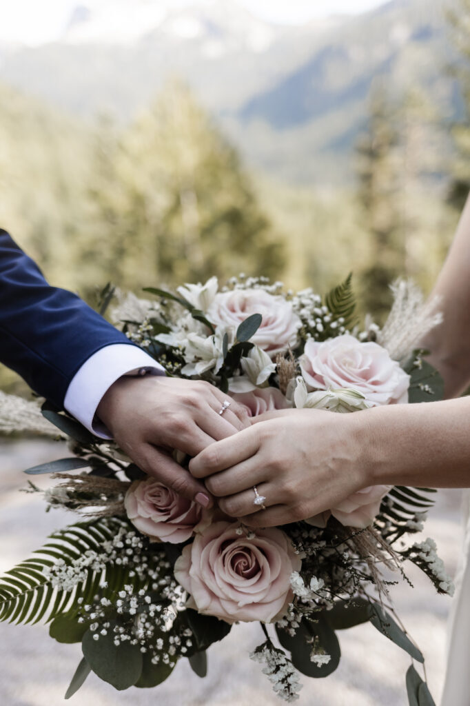 An up-close shot from an LGBTQ+ wedding at the Sea to Sky Gondola in Vancouver. The image consists of the couple holding hands with their wedding rings on display in front of a beautiful pink bouquet of roses and baby's breath.