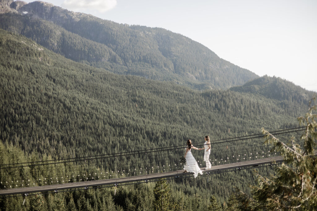 A picturesque scene unfolds as the LGBTQ+ couple, radiating joy, strolls hand in hand along a suspension bridge at the Sea to Sky Gondola in Vancouver. Dressed in a stunning white gown and a white jumpsuit, they traverse the bridge with a backdrop of lush greenery covering the mountains, creating a serene and unforgettable moment.