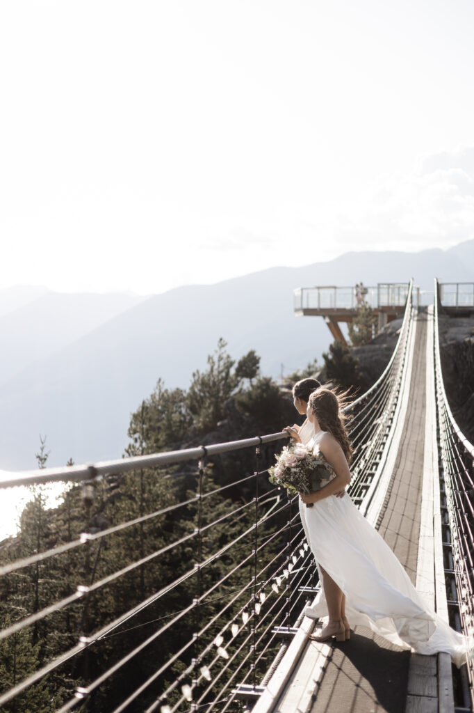 In a serene moment, the LGBTQ+ couple stands side by side on a suspension bridge at the Sea to Sky Gondola in Vancouver. Dressed in a gorgeous white gown and a white jumpsuit, they gaze towards the breathtaking view of mountains and lush greenery, sharing a quiet and intimate moment of reflection and connection against the stunning natural backdrop.