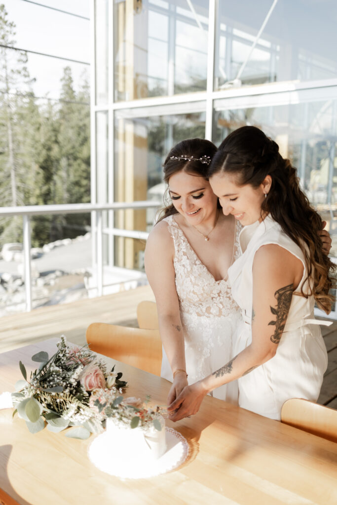 Capturing a joyous moment, the same LGBTQ+ couple, elegantly dressed in a white gown and navy suit, share a tender embrace while cutting their wedding cake. 
