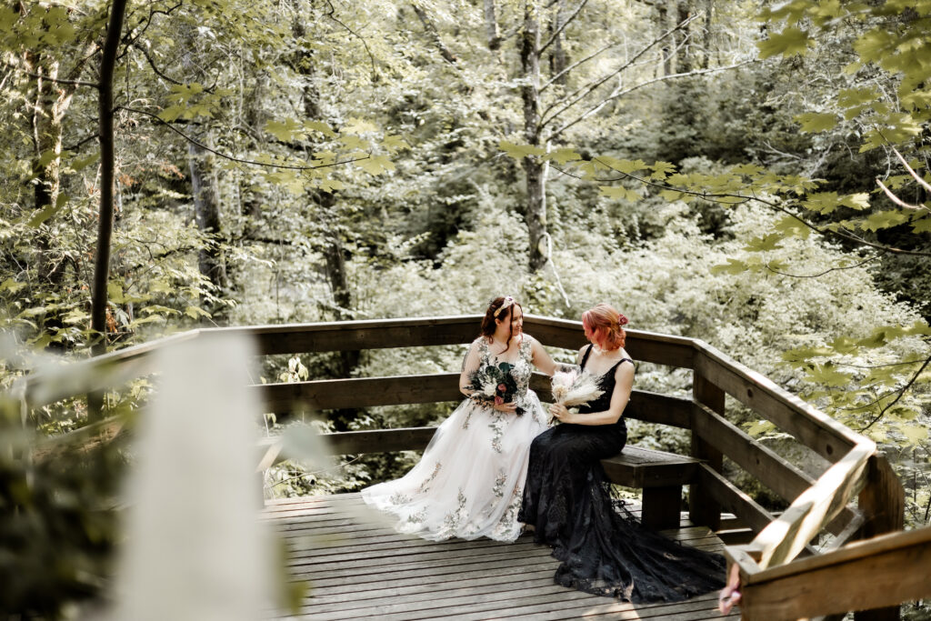 Brides sit beside each other on a wooden bench at their forest elopement in Langley