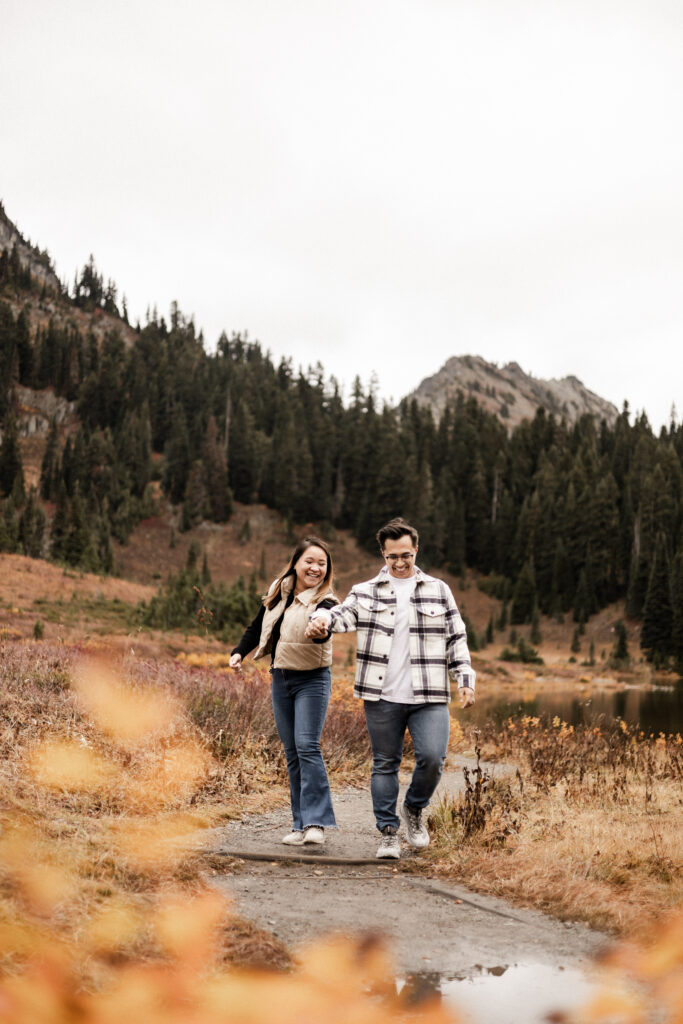 A couple wearing casual clothes hold hands and smile as they walk the pathway around Tipsoo Lake in Mount Raininer National Park. In the background there is a rocky mountain poking out behind a patch of green pine trees.