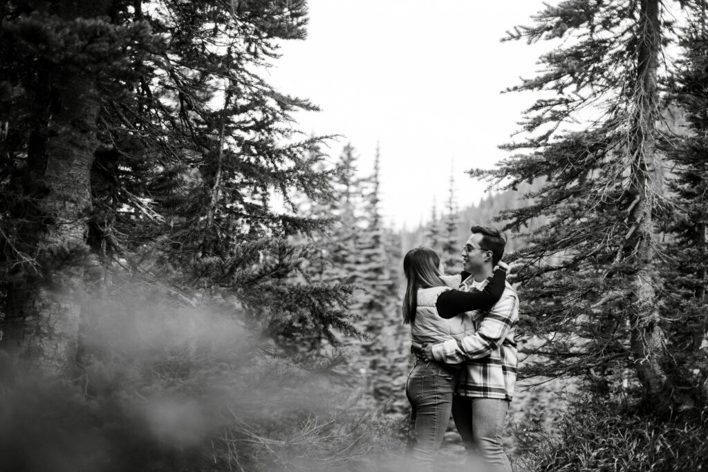 A black and white image of a couple sharing a slow dance among the forests of Mount Rainier Park. The camera is situated behind a tree, placing the branches in the foreground creating perfect framing for the dancing couple.