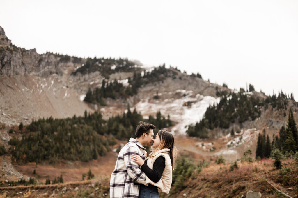 A couple are centred in the image, touching noses and arms wrapped around each other. In the background is a large mountain, partially barren with rocks and patches of green pine trees scattered over the surface.