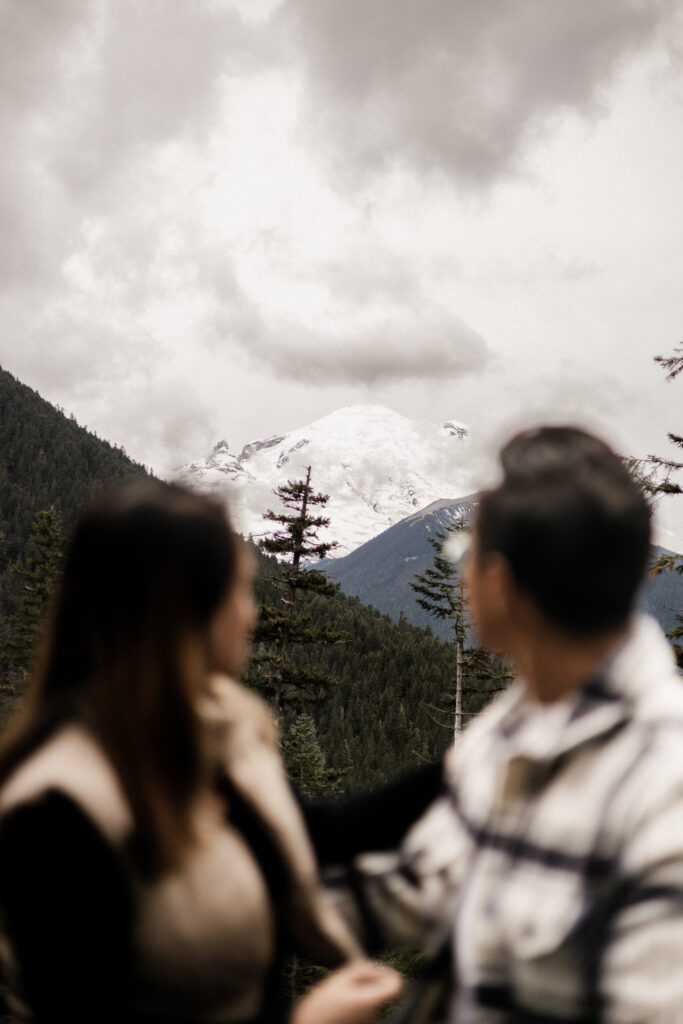 The image changes focus from the couple, who are sitting beside each other looking out, to Mount Rainier. The couple admire the majestic beauty of the mountain, which is covered in snow and partially hidden behind a patch of clouds.
