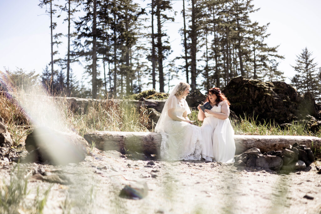 The brides sit together on a log at Big Beach, Ucluelet reading their vows to each other from vow books.