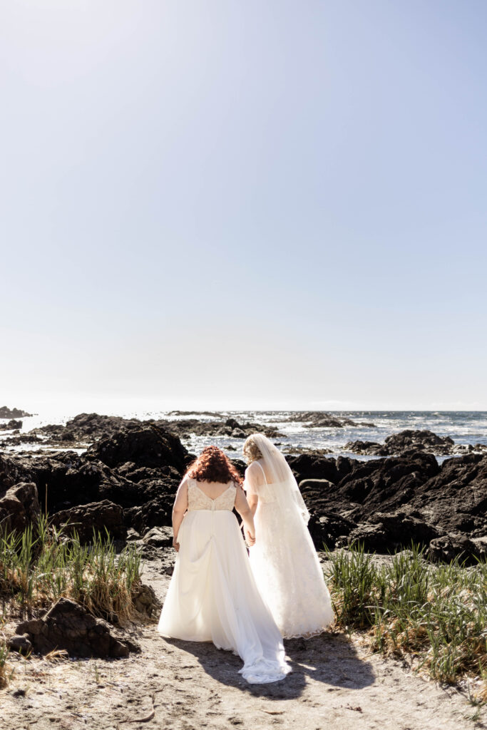 The brides hold hands as they walk together down towards the black rocks on the beach at their Black Rock Oceanfront Resort wedding.