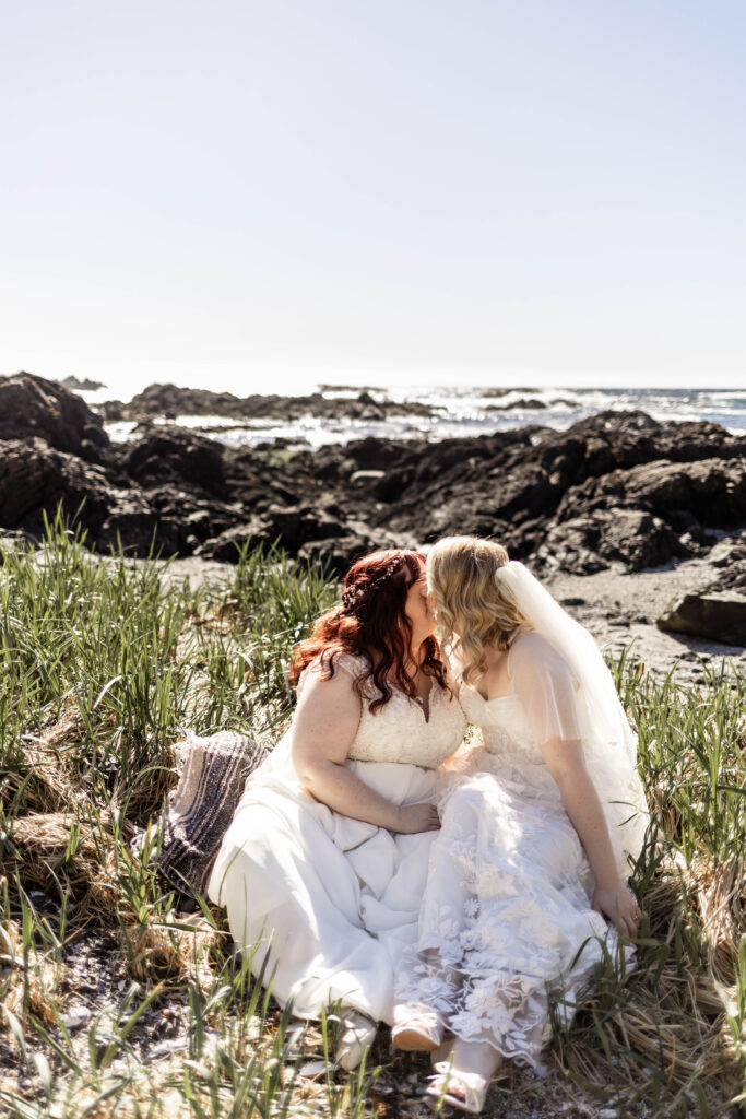 The brides share a kiss as they sit among long grass on the beach at their Black Rock Oceanfront Resort wedding.