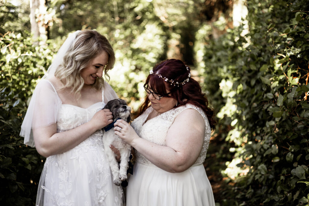 Two brides pose with their small dog in the forest at their Black Rock Oceanfront Resort wedding. They both admire him and give him a pet.