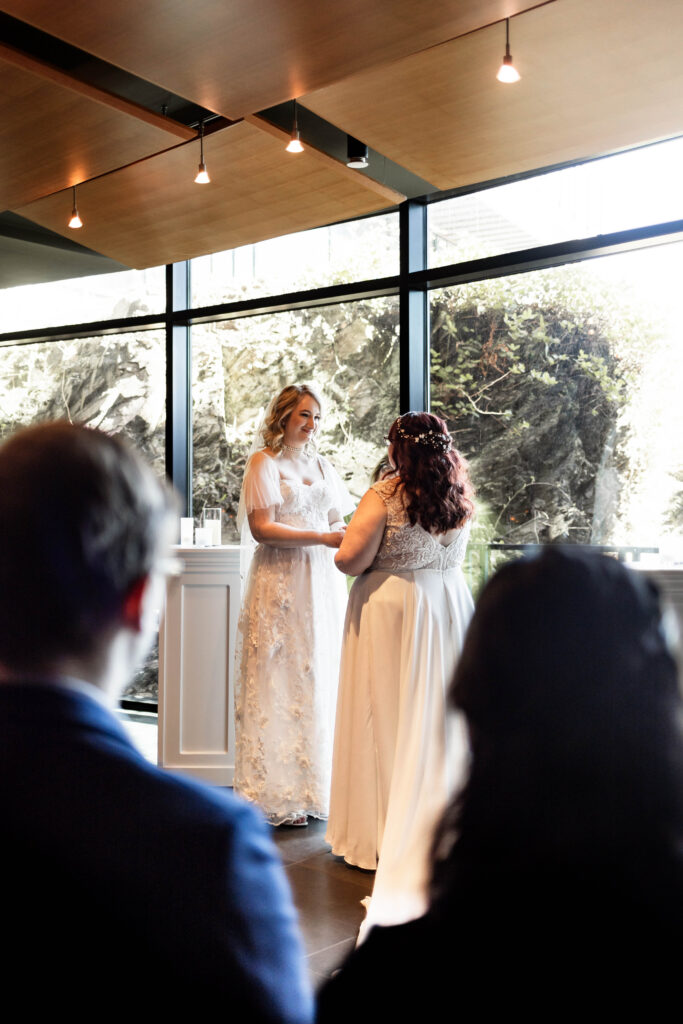 From the point of view of their wedding guests, two brides hold hands at the altar of their ceremony at their Black Rock Oceanfront Resort wedding.