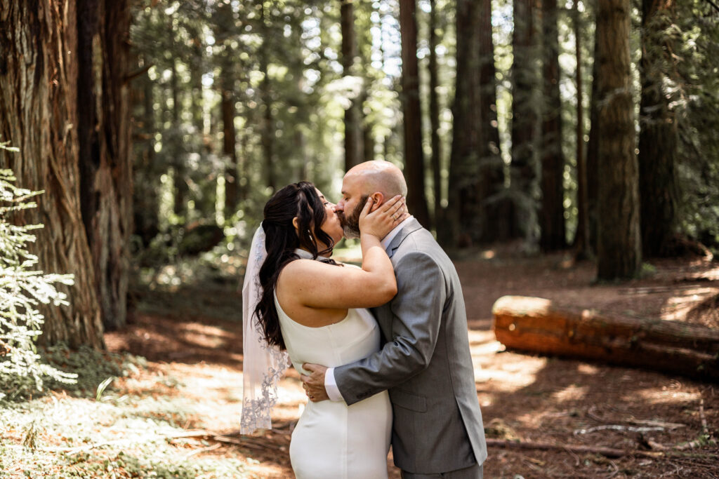 A couple share a first kiss as a married couple standing amongst redwood trees.