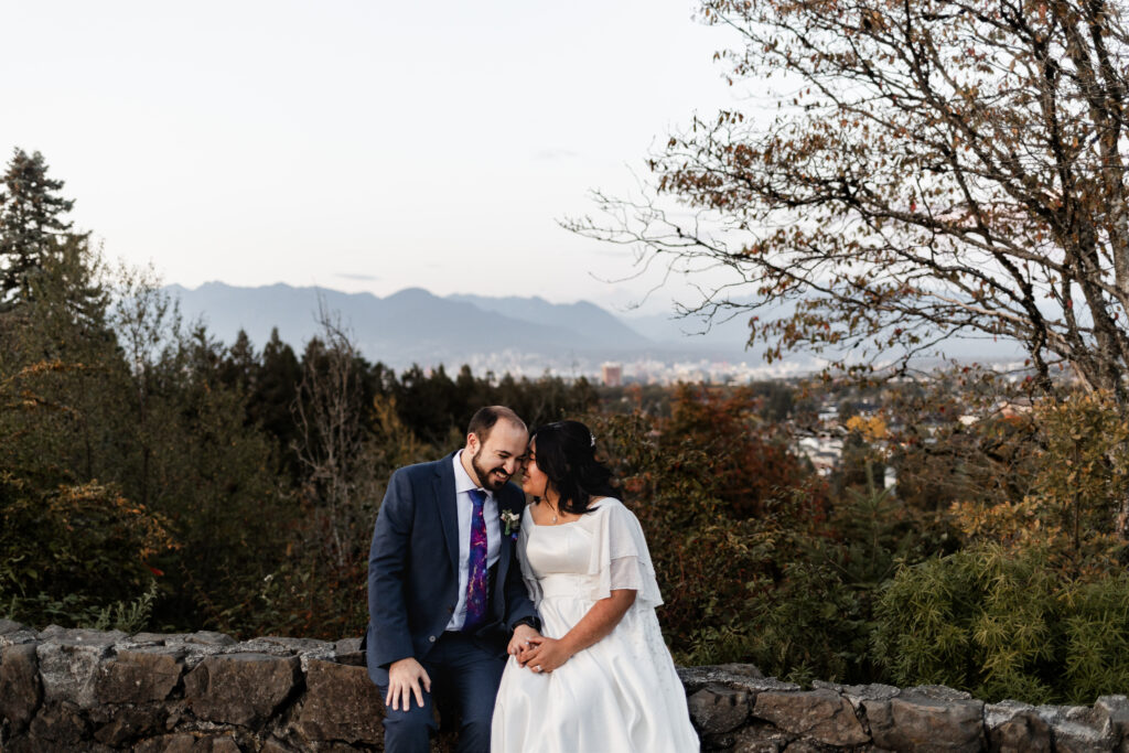 A bride and groom sit together on a low rock wall in front of a view of Vancouver at their traditional wedding.