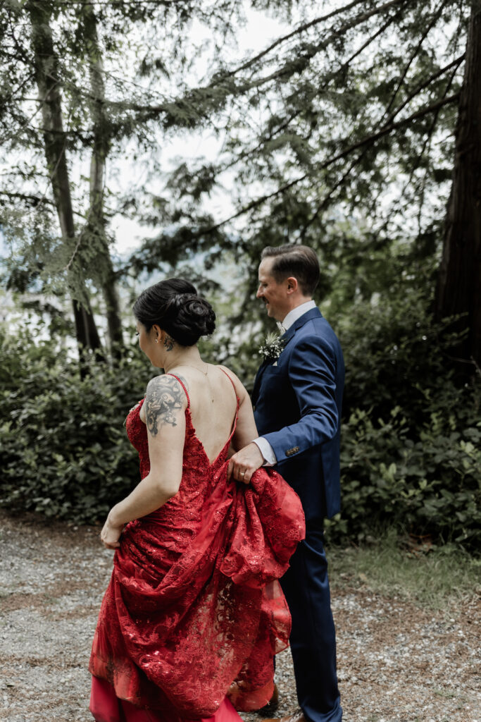 The groom holds the train of the bride's red dress as they walk at their Gibsons Island summer camp wedding at Camp Elphinstone