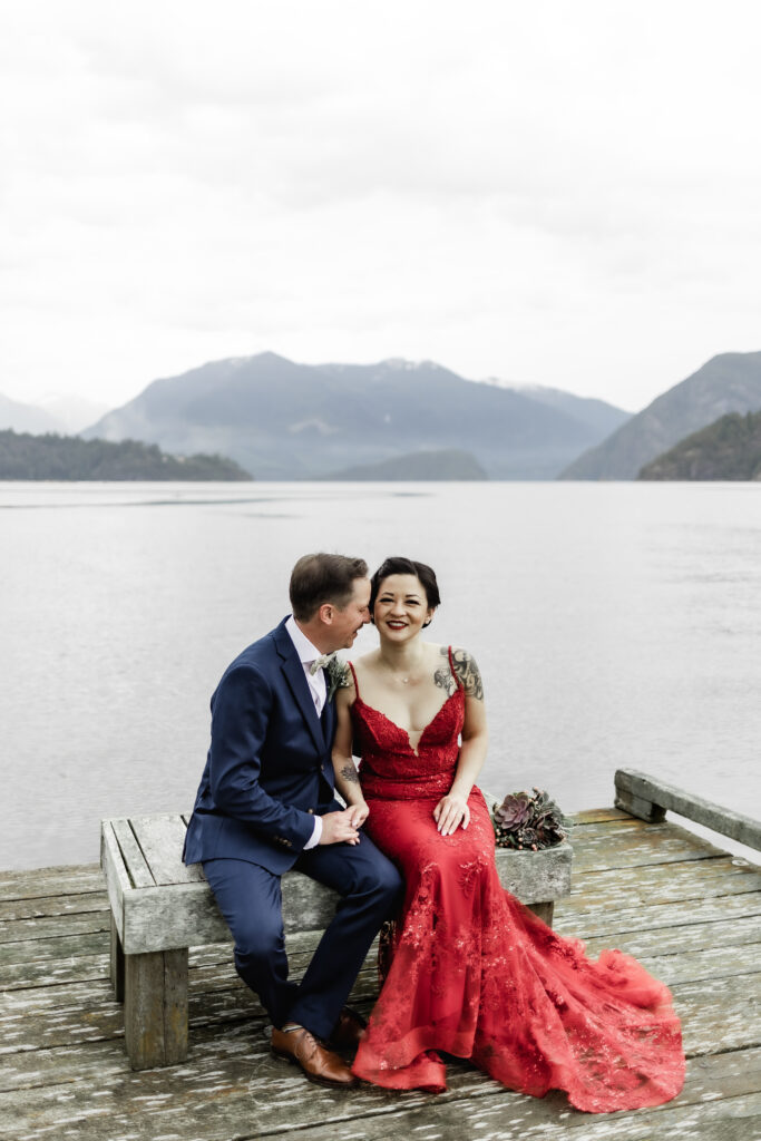 The bride and groom sit next to each other on a bench in front of the ocean and mountain view at their Gibsons Island summer camp wedding at Camp Elphinstone