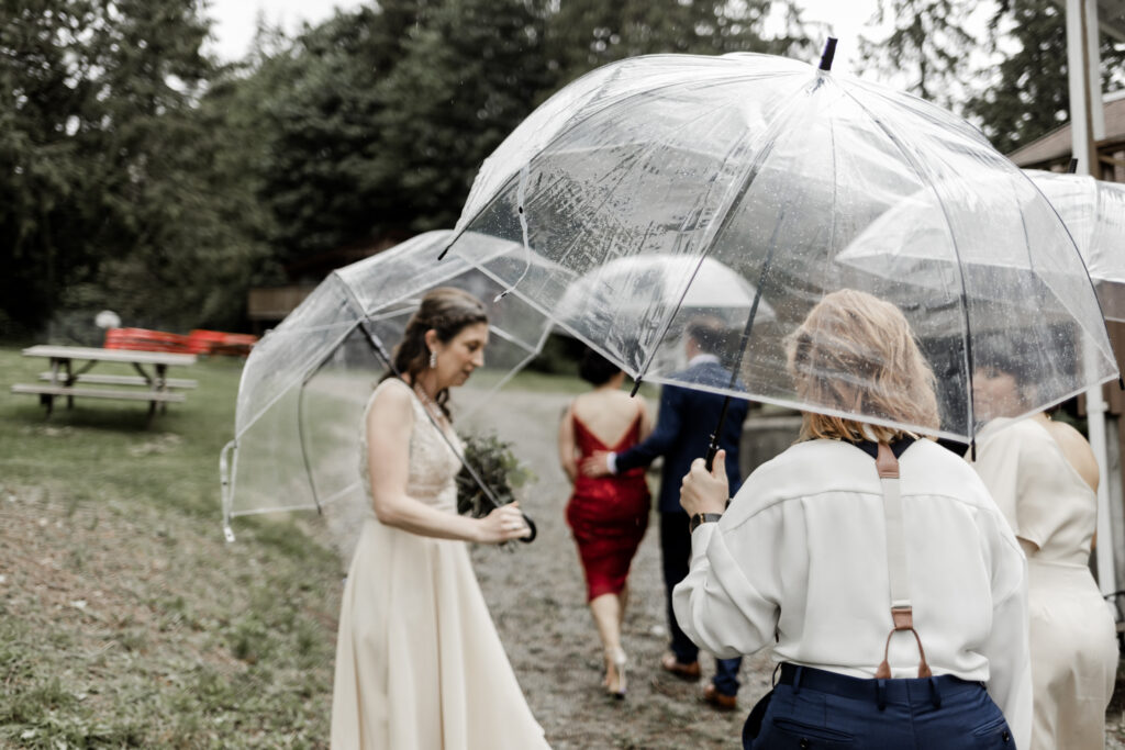 The bridal party walks with clear umbrellas at this Gibsons Island summer camp wedding at Camp Elphinstone