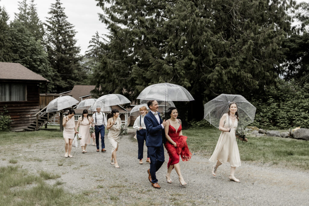 The bridal party walks with clear umbrellas at this Gibsons Island summer camp wedding at Camp Elphinstone