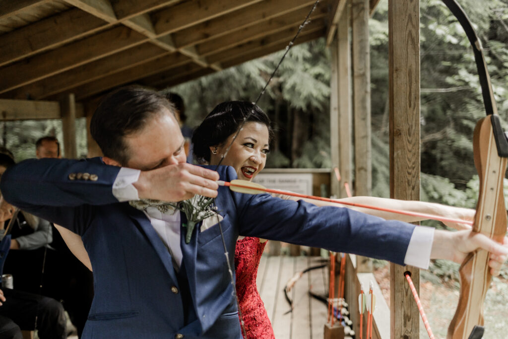 The bride helps her groom aim his bow at the target at this Gibsons Island summer camp wedding at Camp Elphinstone