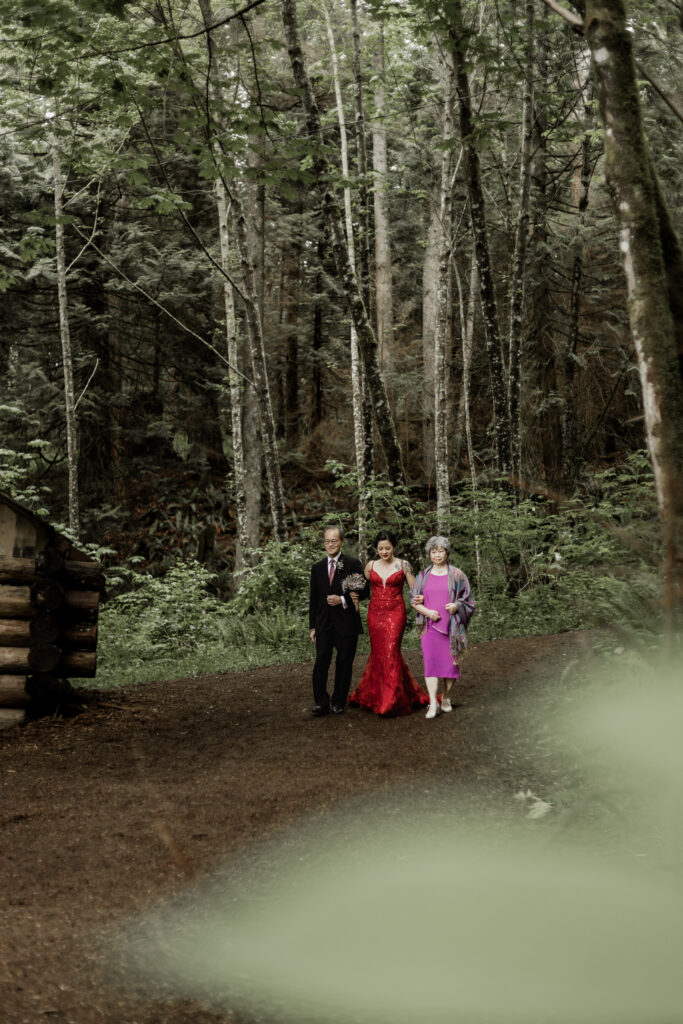 The bride walks arm in arm with her parents down the aisle at this Gibsons Island summer camp wedding at Camp Elphinstone