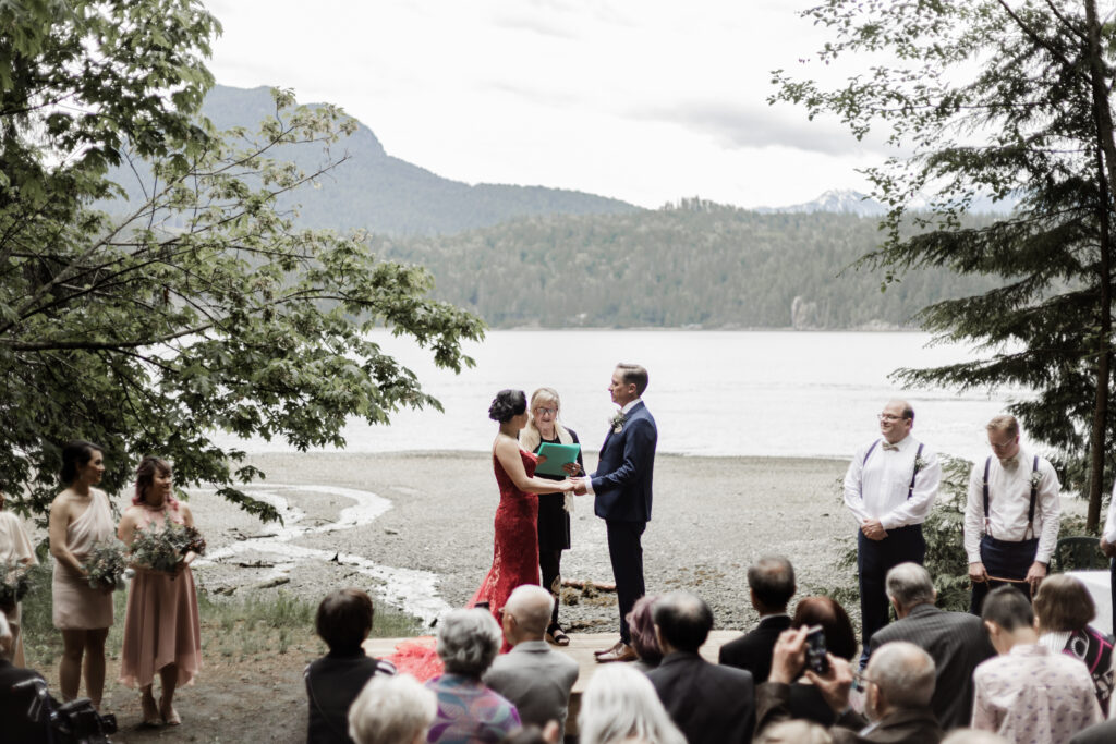 The couples hold hands at the altar in front of an ocean and mountain view at this Gibsons Island summer camp wedding at Camp Elphinstone