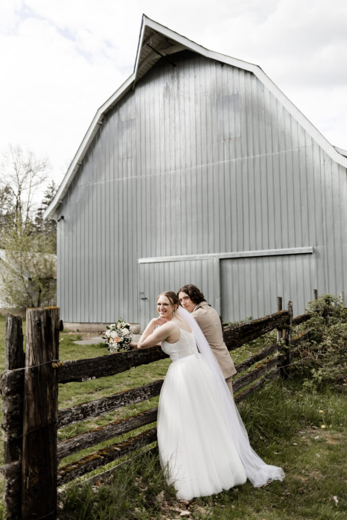 Bride and groom lean on a fence and look at the blue barn at this Estate 248 wedding in Langley, British Columbia